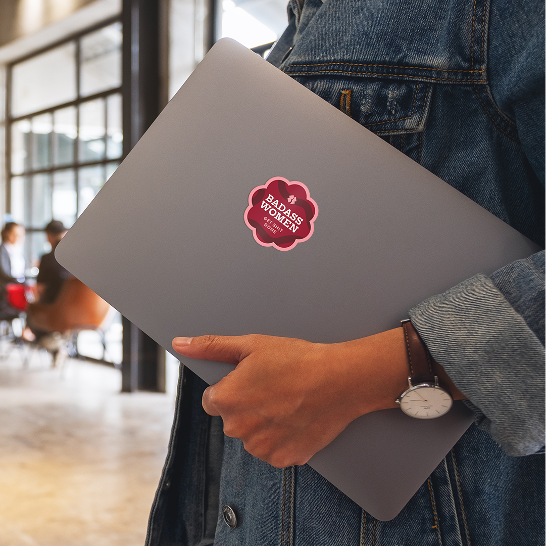 Person holding a gray laptop with a 'Badass Women' sticker in an indoor setting