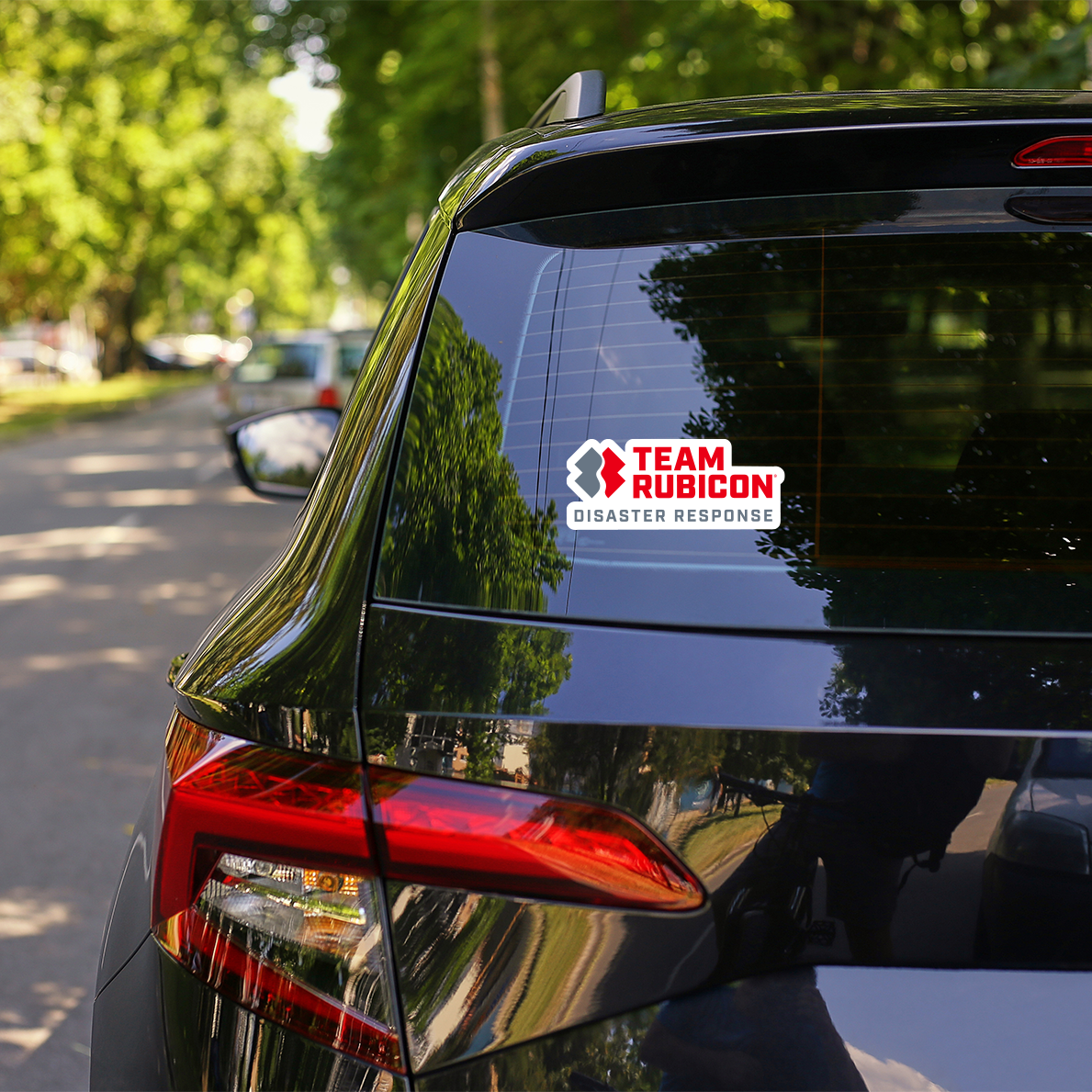 A Team Rubicon Disaster Response decal applied to the rear window of a black SUV, with a blurred background of trees and a road.