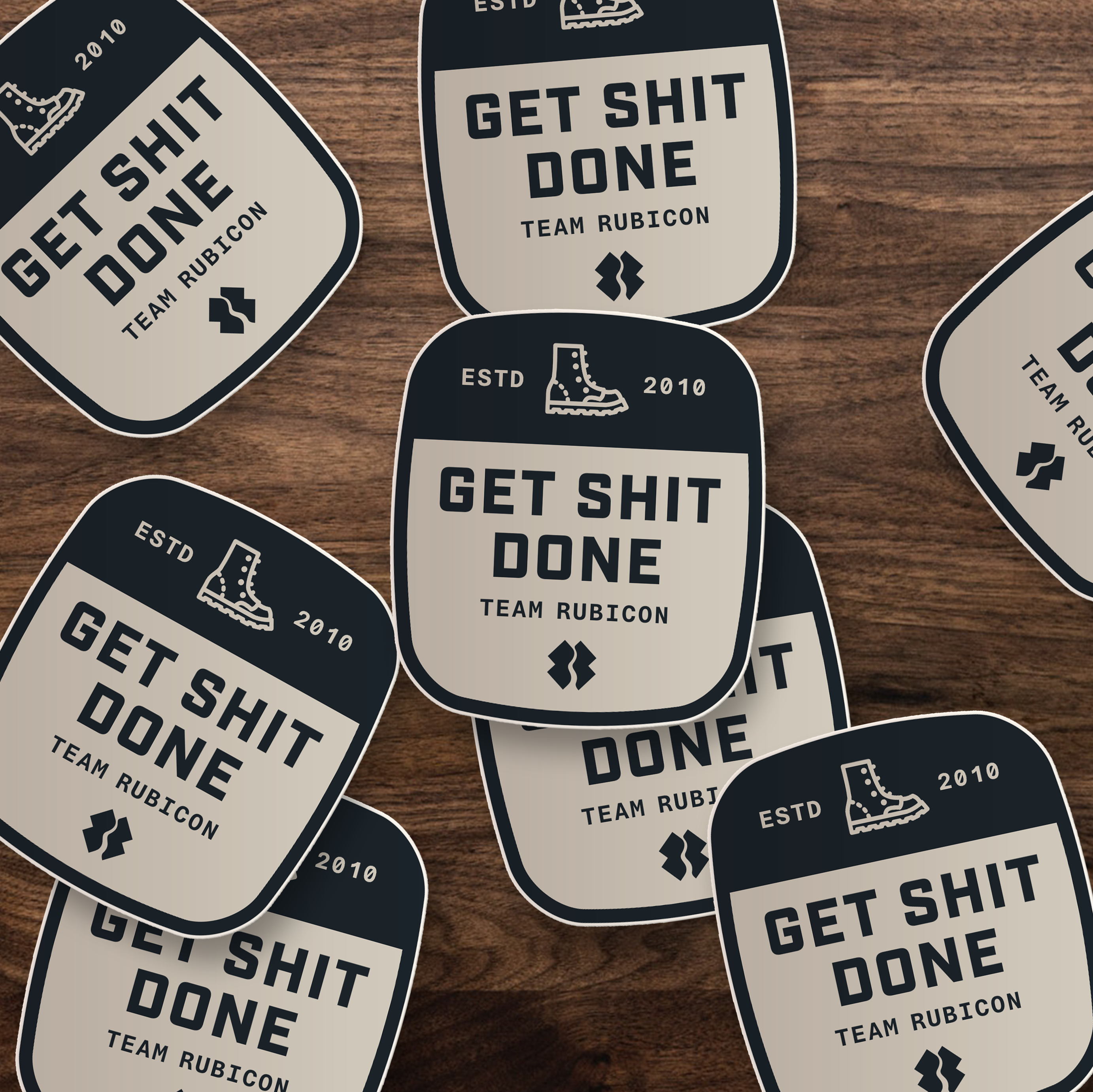 Several die-cut stickers with rounded corners scattered on a wooden surface. Each sticker has a dark navy border and a warm cream center with the text 'GET SHIT DONE TEAM RUBICON' and small boot icons.
