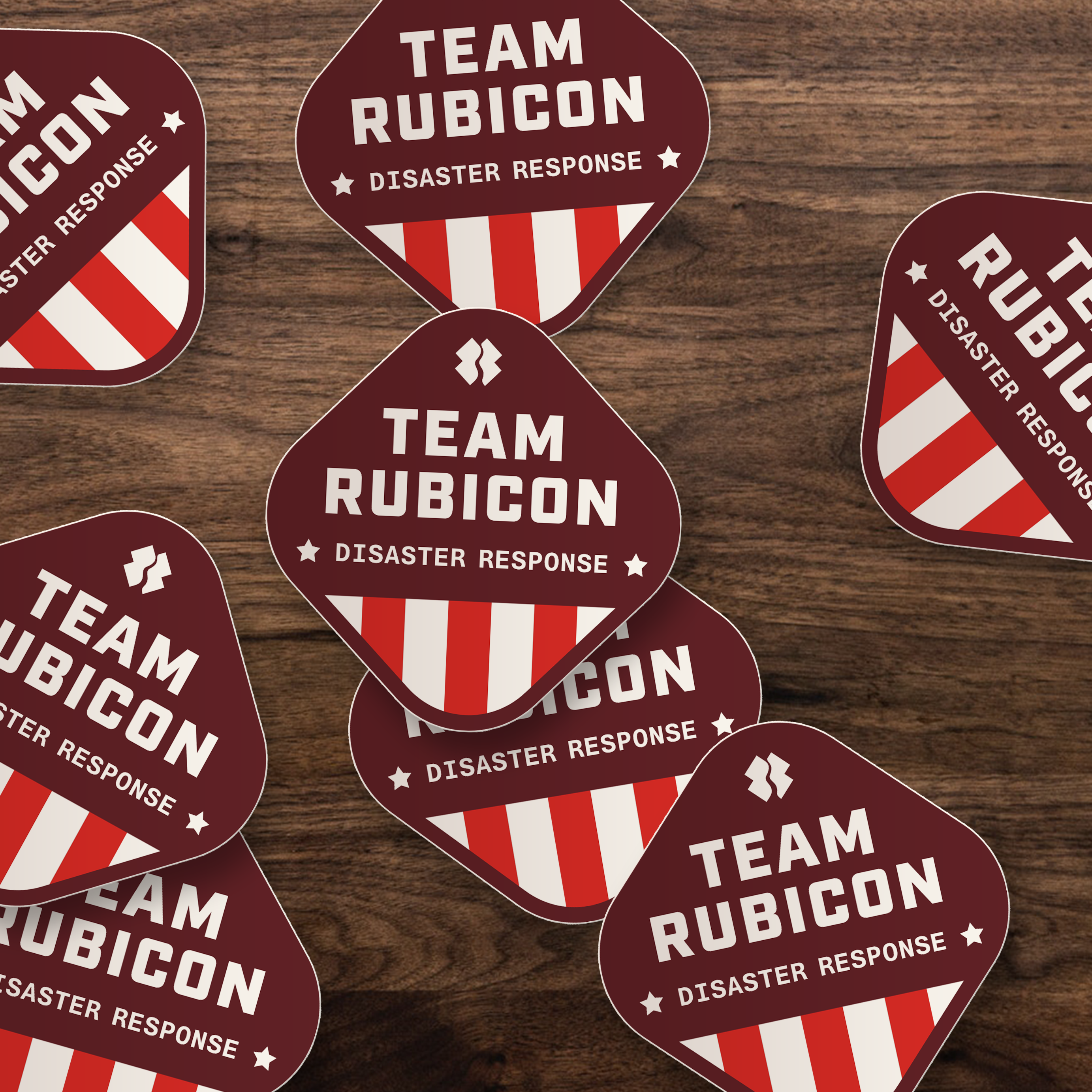 Several die-cut stickers with rounded shield-like shapes arranged on a dark wood surface. Each sticker is dark red with white text reading "TEAM RUBICON" and "DISASTER RESPONSE," plus diagonal red-and-white stripes.