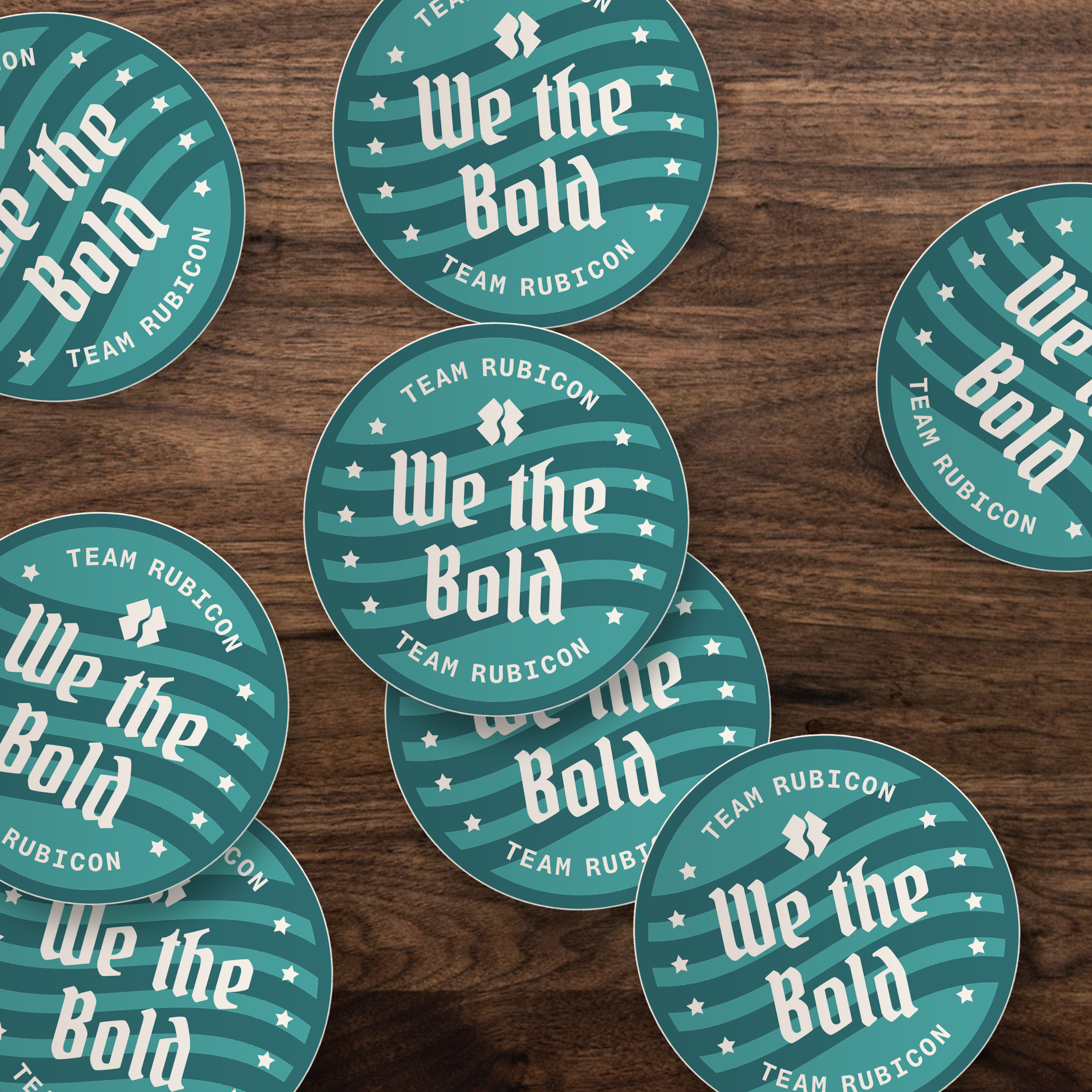 Several round teal and white stickers pieces printed with the text 'We the Bold' and 'TEAM RUBICON' are arranged on a dark wood surface.